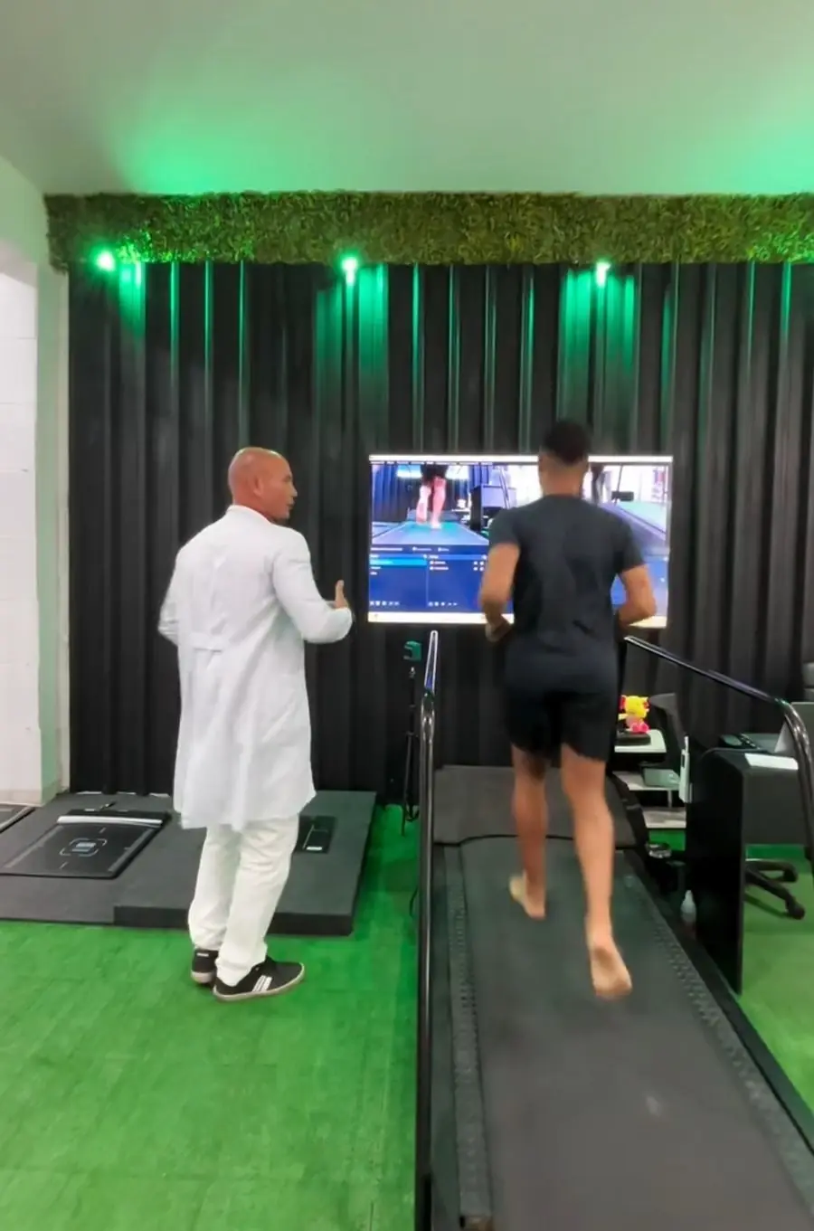 Léo ao lado de um paciente na esteira, os dois olhando a análise da caminhada na TV