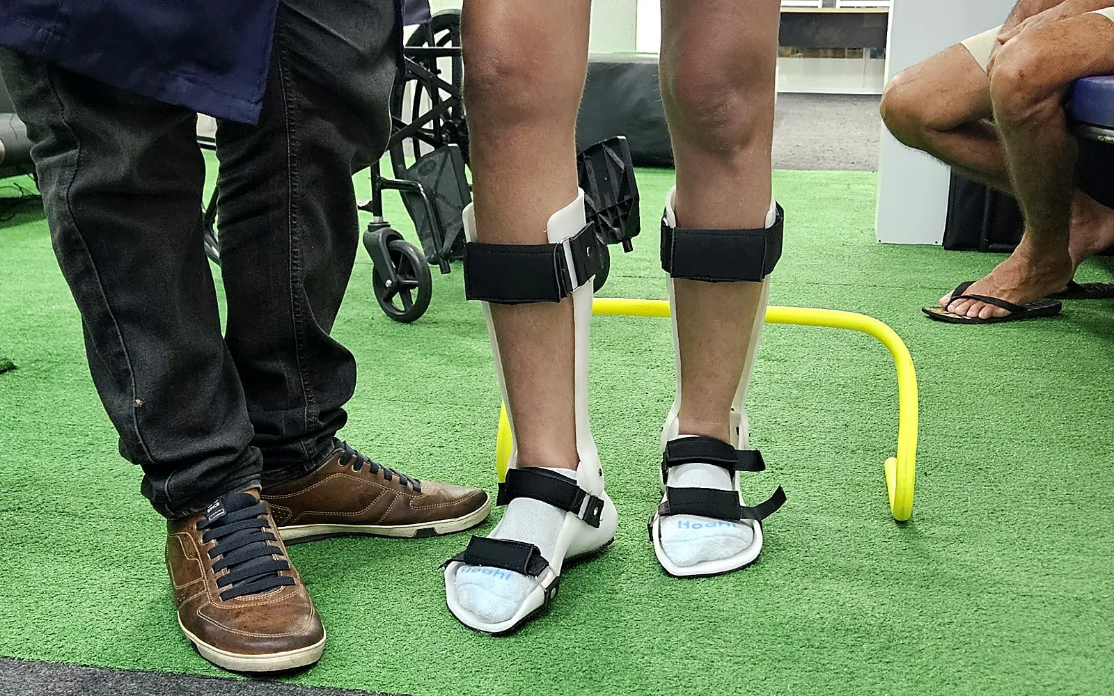 Paciente em pé usando órteses de tornozelo e pé, acompanhada pelo fisioterapeuta